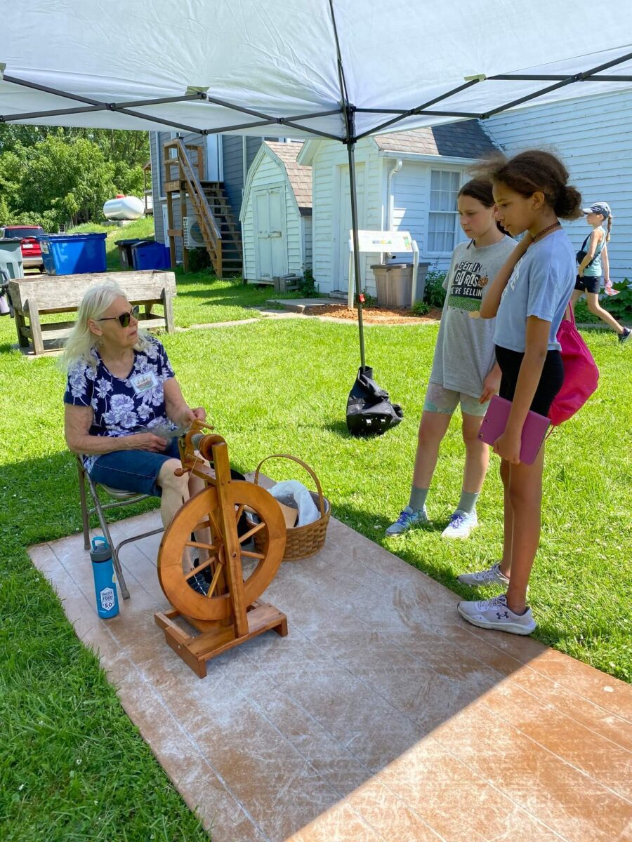 An older woman demonstrates a spinning wheel to two girls under a canopy on a sunny lawn.