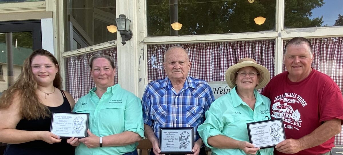 Five people stand outside a building, four holding framed awards, smiling for a group photo.