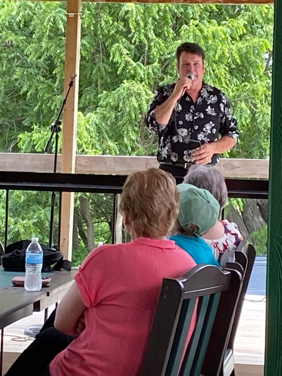 Man in a floral shirt sings into a microphone on a stage before an audience seated outdoors.