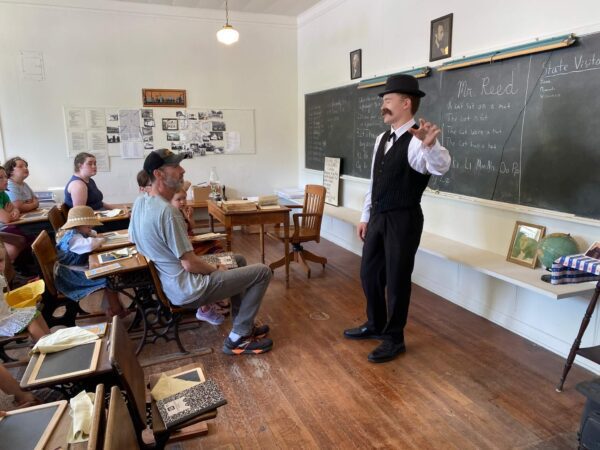 A man in vintage clothes teaches a group in an old-fashioned classroom with desks and a chalkboard.
