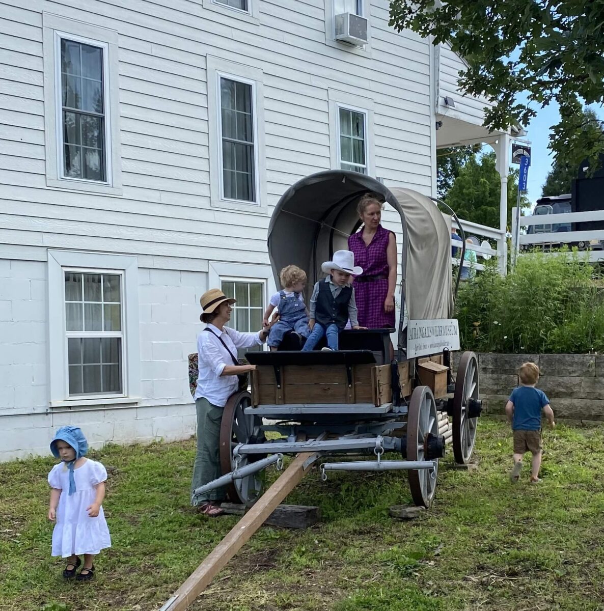 Several children and two adults play on and around a covered wagon beside a large white building.