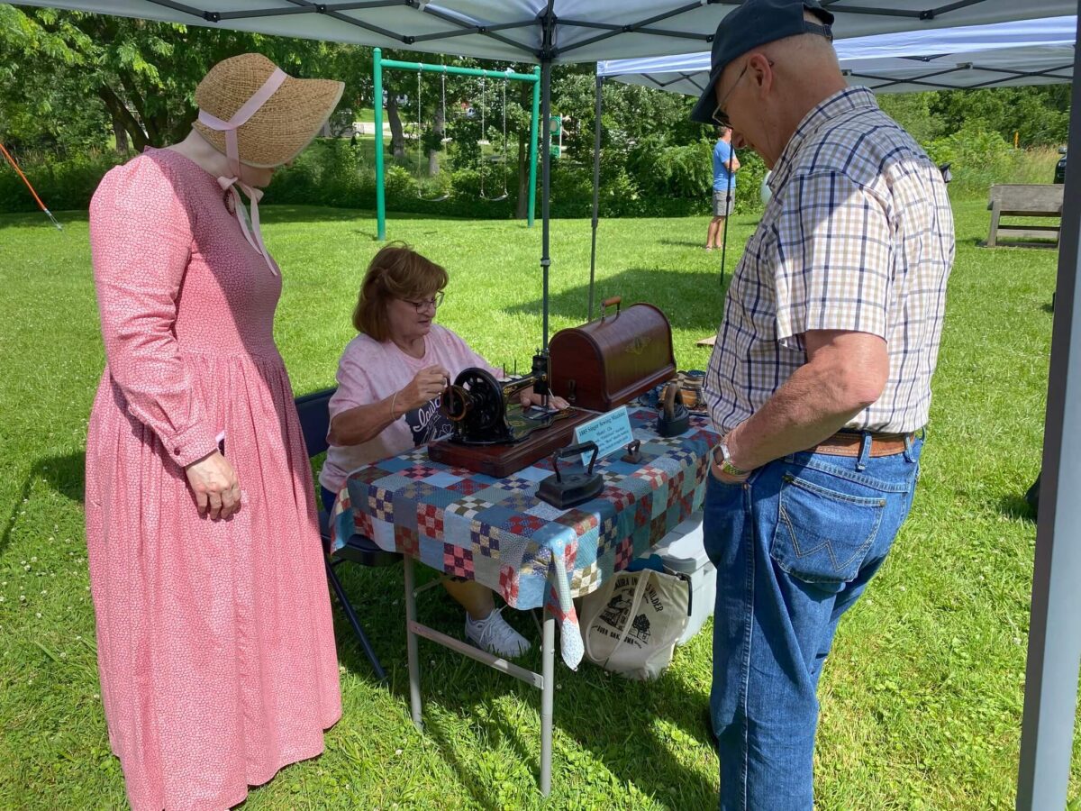 Three people watch a woman using a vintage sewing machine at an outdoor event under a tent.