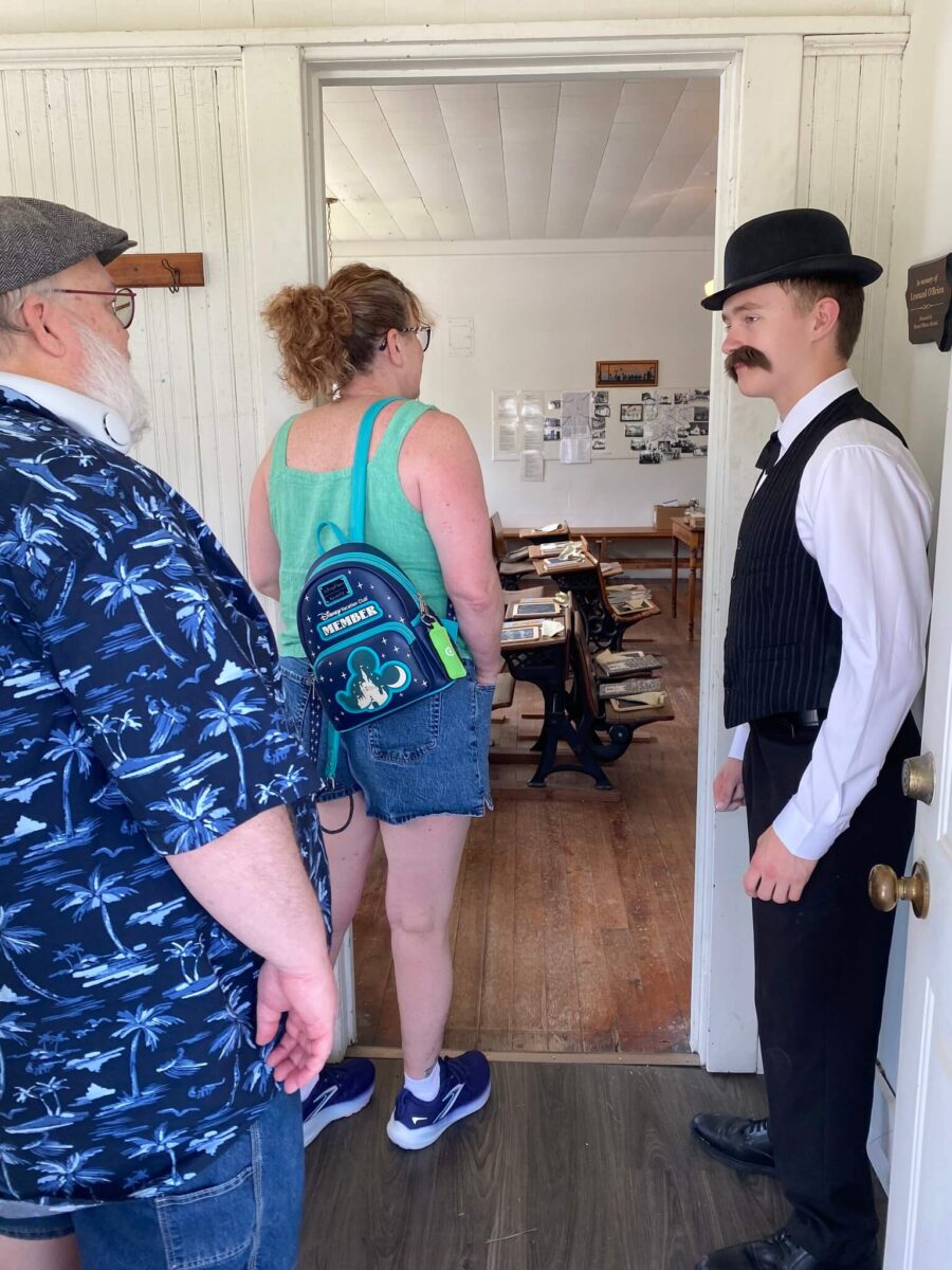 Two visitors enter a historical room as a man in period costume stands by the door, welcoming them.
