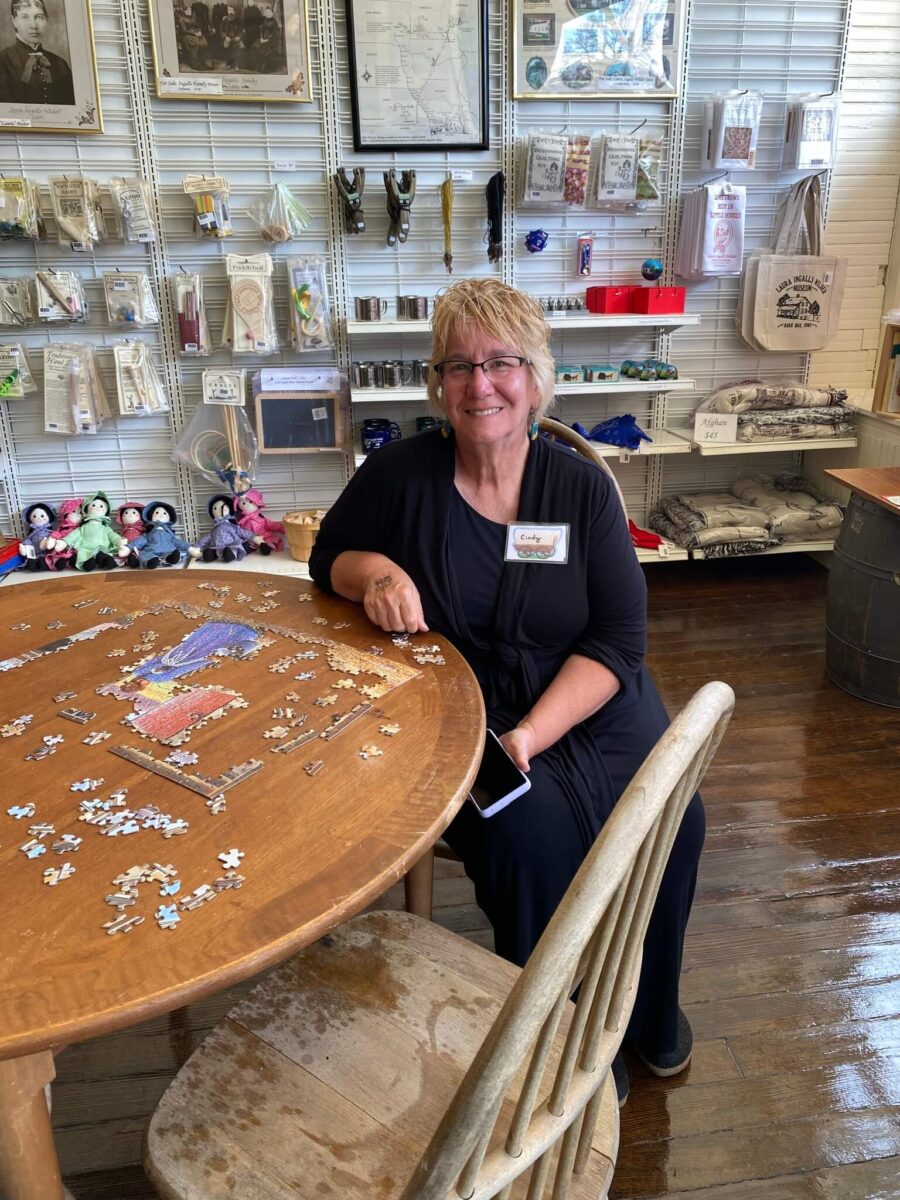 Smiling woman sits at a round table working on a jigsaw puzzle in a gift shop with shelves of souvenirs.