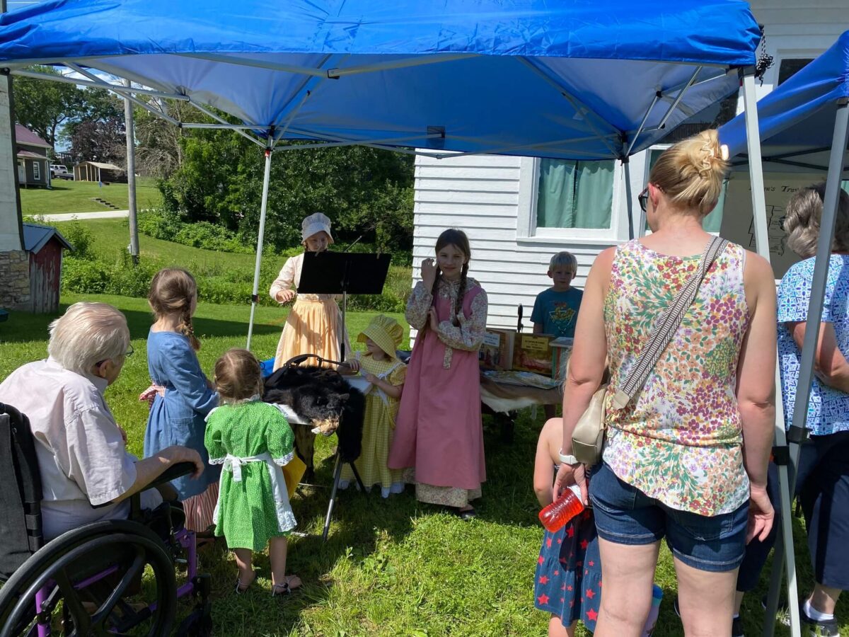 People gather outdoors under tents, some in historical costumes, at a community event on a sunny day.