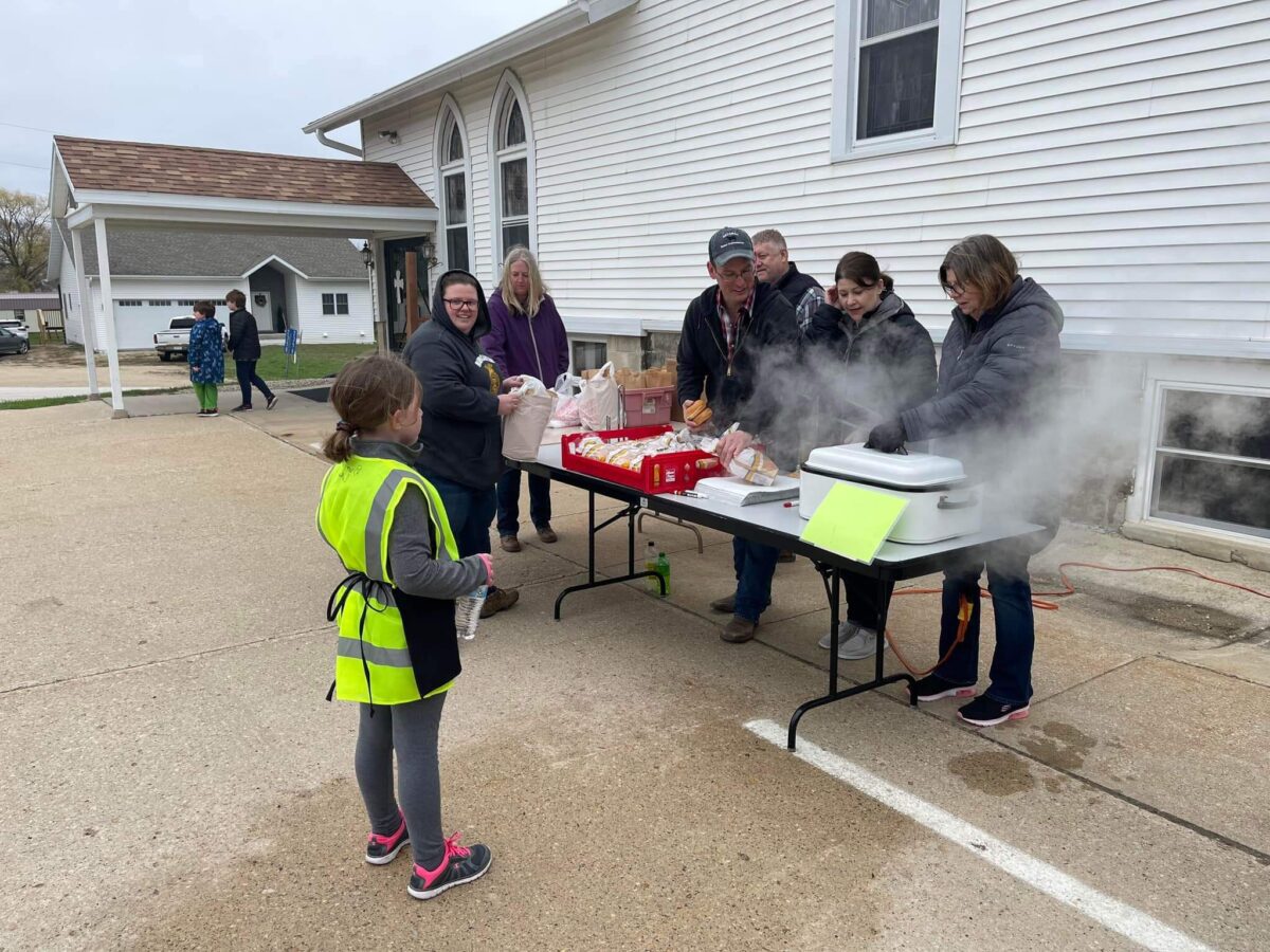 Several people serve food at a table outside a building, while a child in a yellow vest stands nearby.