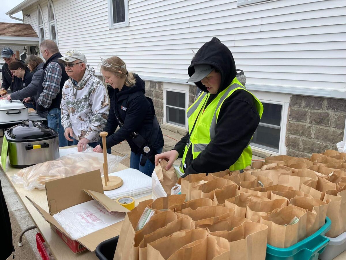 People assemble and pack paper bag lunches at an outdoor table near a white building.