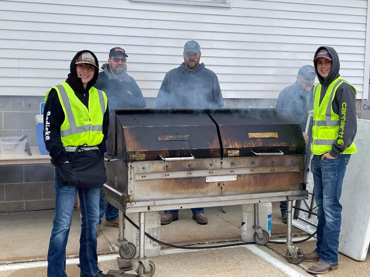 Four people in yellow safety vests stand around a smoking outdoor grill in front of a white building.