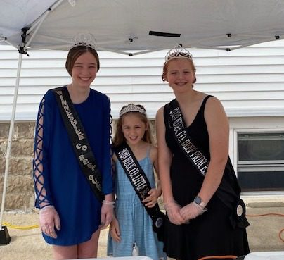 Three girls wearing tiaras and sashes smile under a tent, posing together at an outdoor event.
