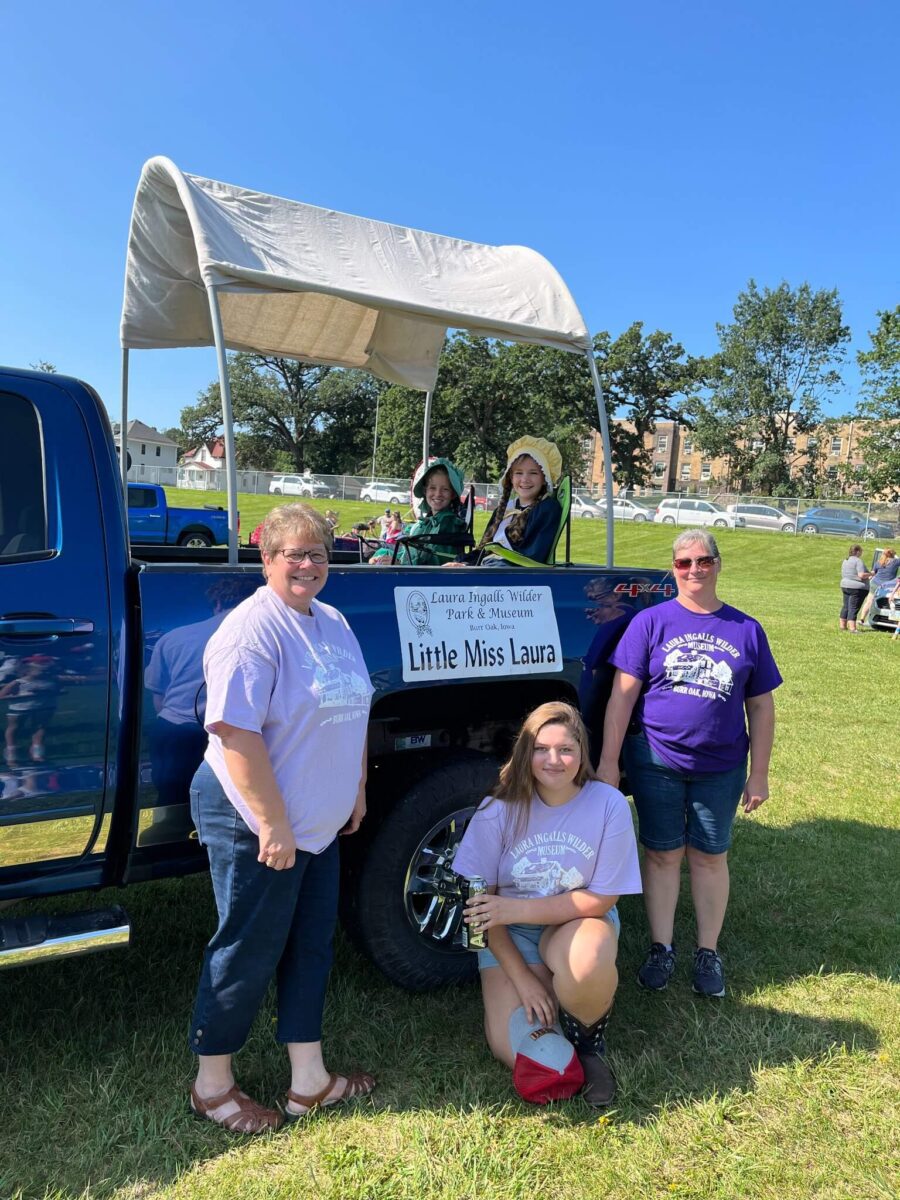 Four women pose by a blue truck with a sign reading 