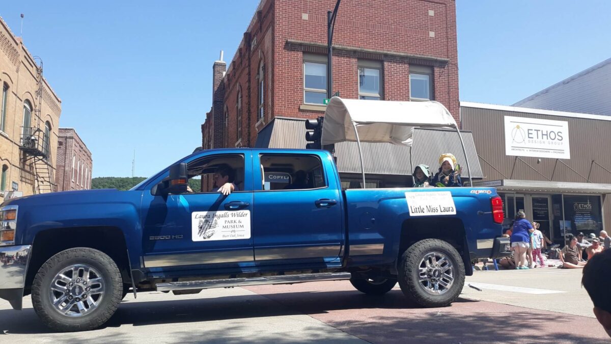 A blue pickup truck pulls a covered wagon in a parade, with people riding in both, on a sunny day downtown.