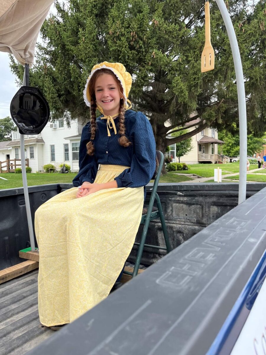 Young girl in pioneer-style dress and bonnet sits on a chair in a wagon, smiling, with houses in the background.