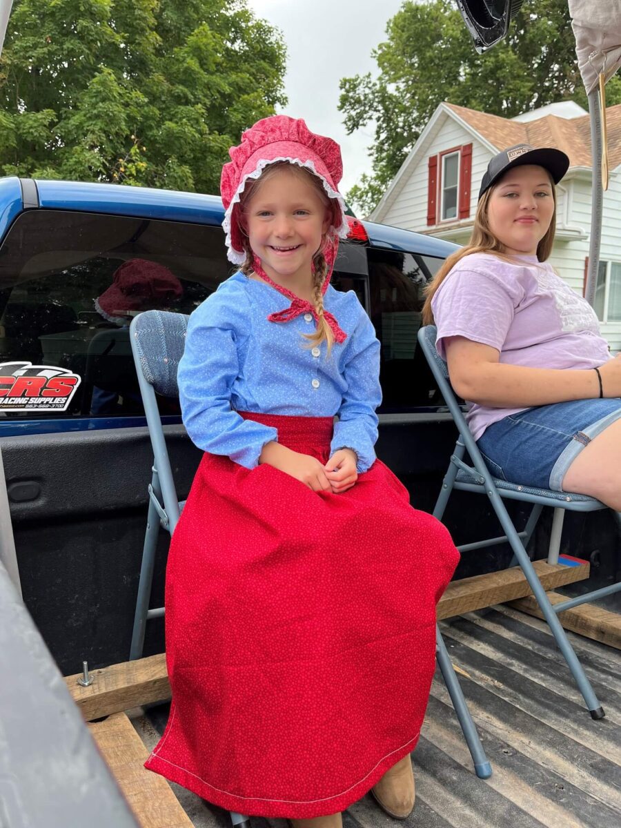 Smiling girl in a bonnet and pioneer dress sits on a chair; another girl in casual clothes sits behind her.