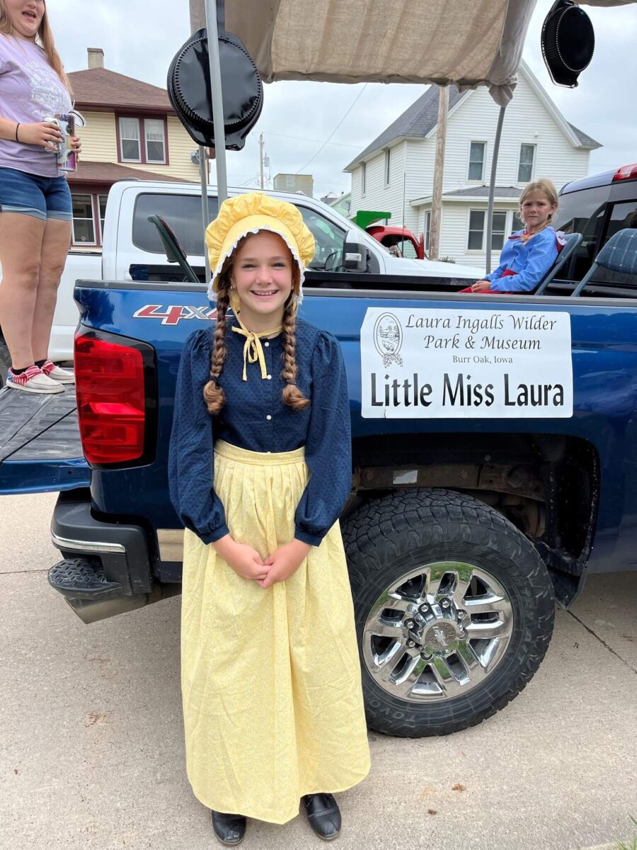 Smiling girl in a bonnet and pioneer dress stands by a truck with a 