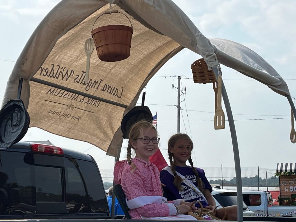 Two girls in costumes sit in a covered wagon parade float with baskets and utensils hanging overhead.