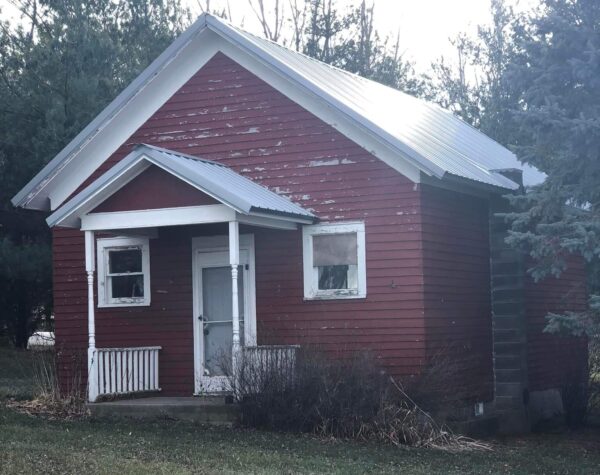 Small, weathered red cabin with white trim, surrounded by grass and trees on a sunny day.