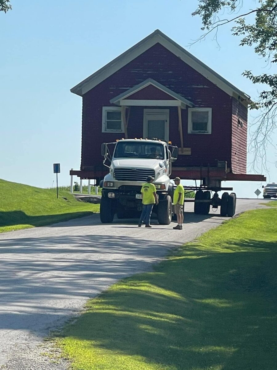 A small house is being transported on a truck down a rural road, guided by three workers in yellow vests.
