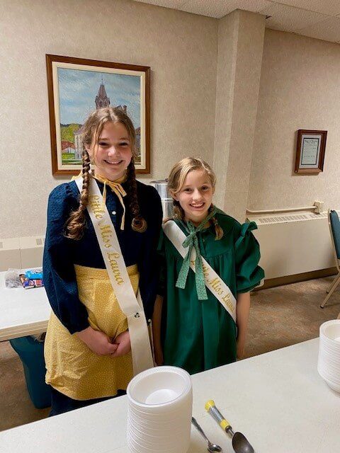Two girls with sashes and braids stand behind a table with plates, smiling in a room with a framed picture.
