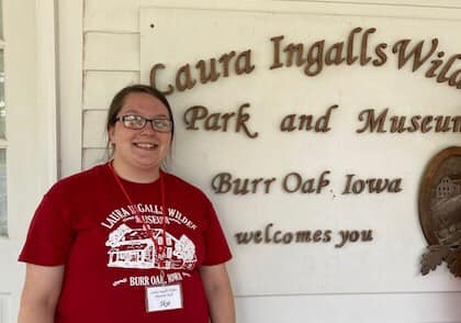 Person in a red shirt standing and smiling beside a Laura Ingalls Wilder Museum sign in Burr Oak, Iowa.