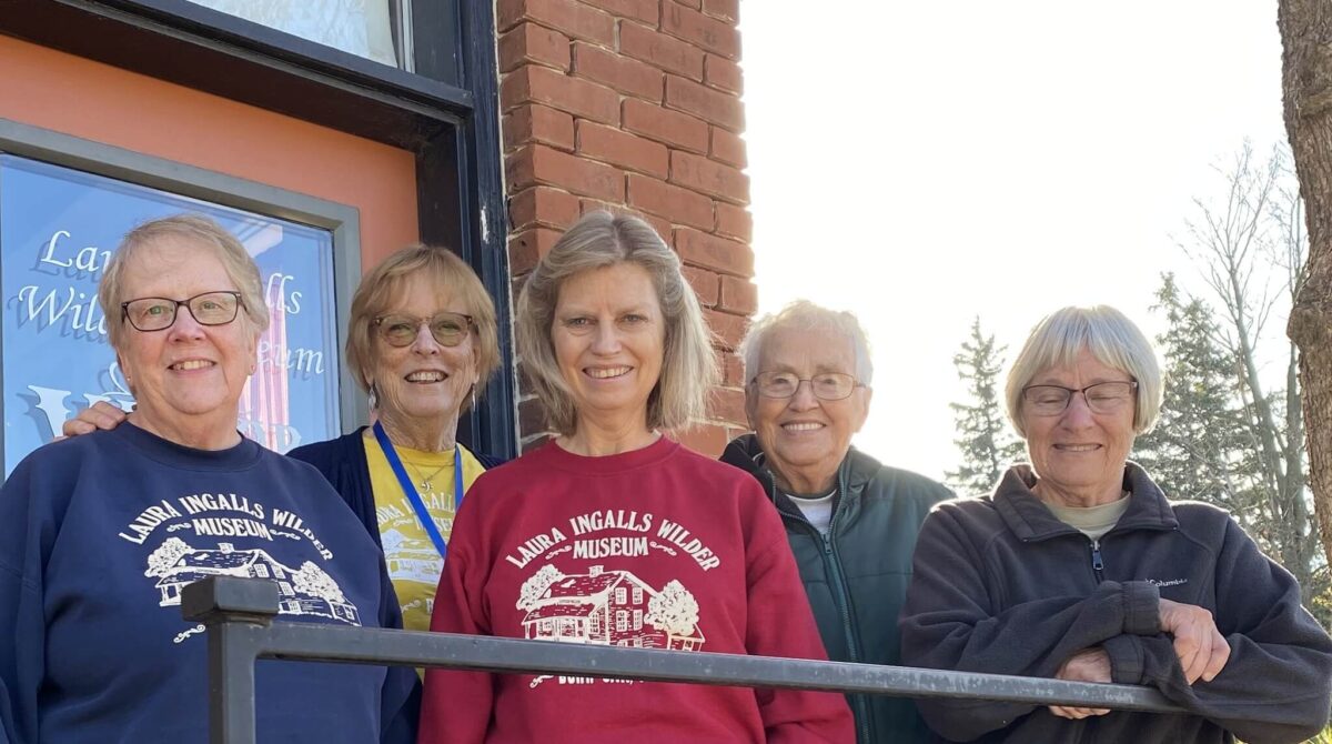 Five smiling women stand together outside a brick building, some wearing Laura Ingalls Wilder Museum shirts.