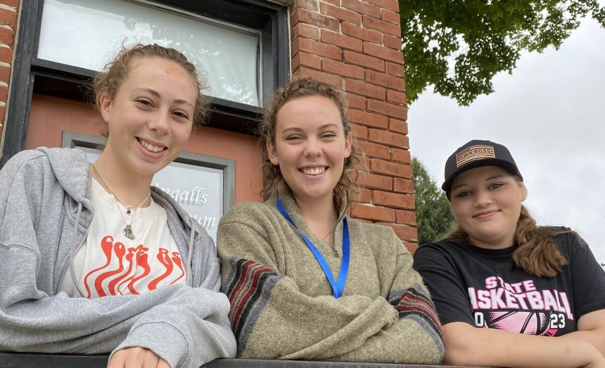 Three young women smiling outside near a brick building, with trees and a cloudy sky in the background.