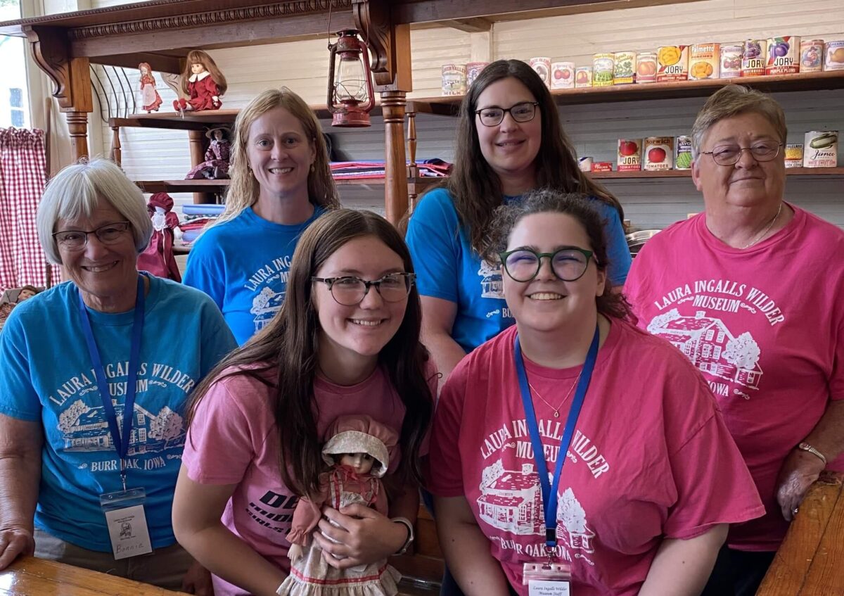 Six smiling women in Laura Ingalls Wilder Museum shirts pose together inside a shop with vintage items on shelves.