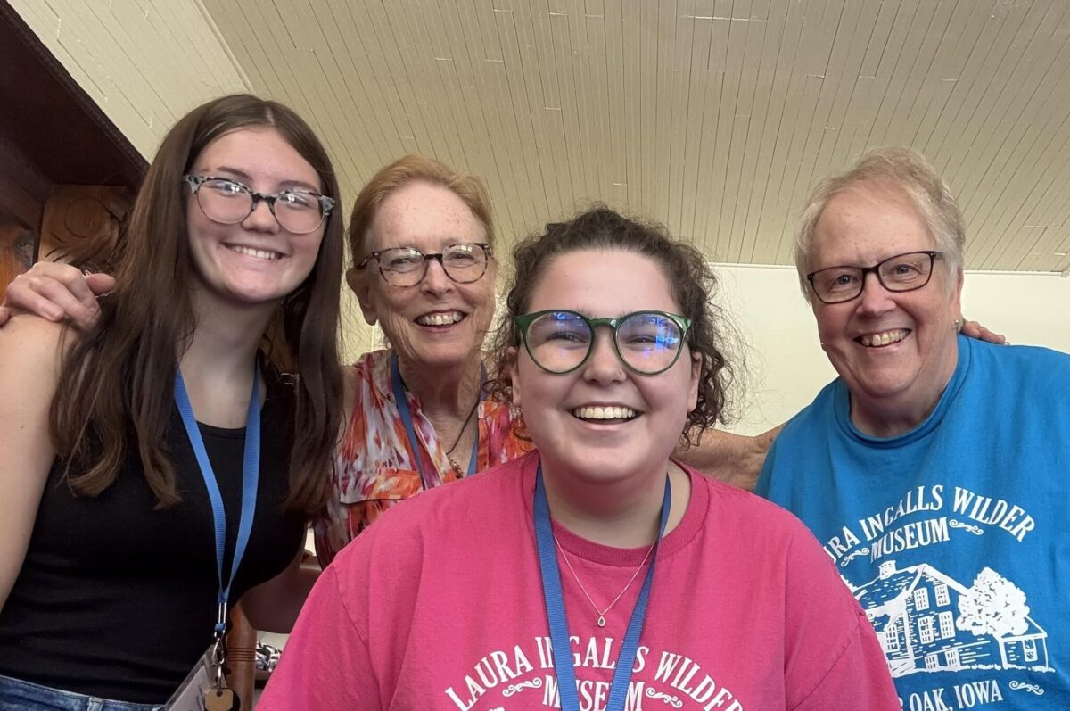 Four people smiling indoors, all wearing glasses and colorful Laura Ingalls Wilder-themed shirts.