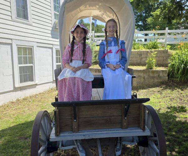Two girls in pioneer costumes sit in a covered wagon outside a white building on a sunny day.
