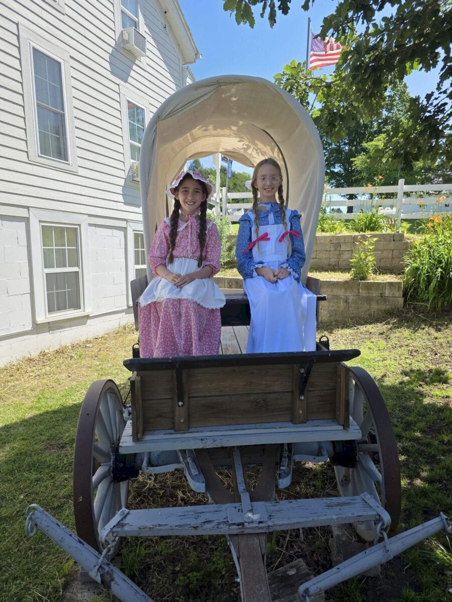 Two girls in pioneer costumes sit in a covered wagon outside a white building on a sunny day.